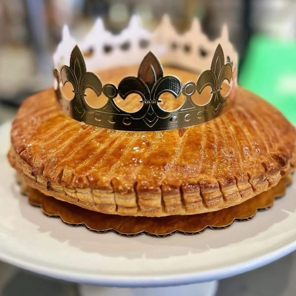 Golden brown Galette des Rois, or King Cake, topped with a silver paper crown on a white cake stand.