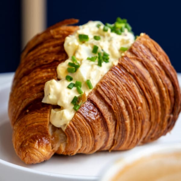 Golden-brown croissant filled with creamy egg salad and topped with fresh green chives on a white plate.