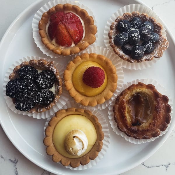 Six assorted fruit tarts, including strawberry, blueberry, blackberry, raspberry, and lemon, on a white plate.