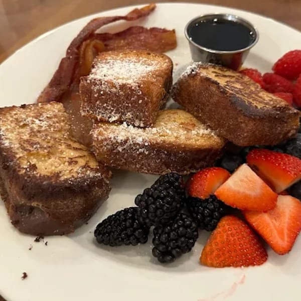 French toast sticks with powdered sugar, bacon, strawberries, blackberries, and syrup on a white plate.