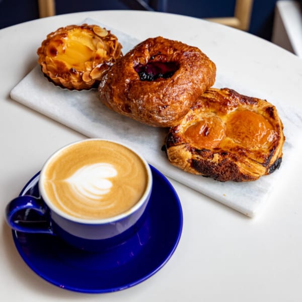 A blue cup of latte with heart-shaped foam art next to three pastries on a marble slab.