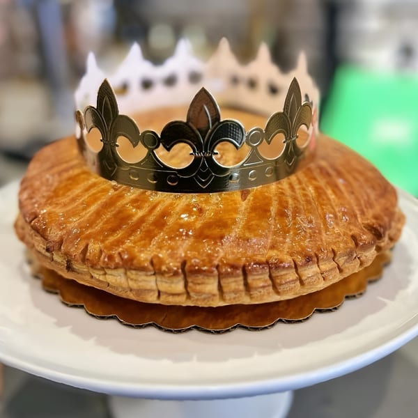 Golden-brown Galette des Rois pastry with a silver paper crown on a white cake stand.