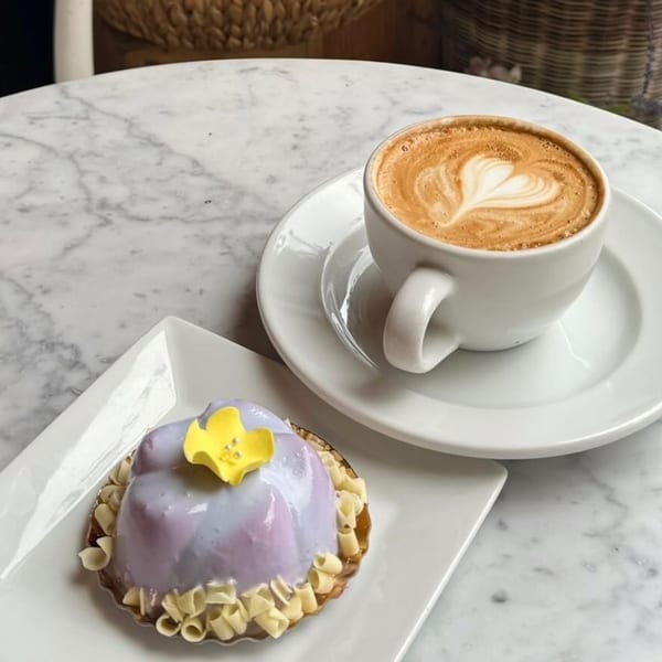 A purple glazed pastry with a yellow flower and white chocolate curls next to a latte with heart art.