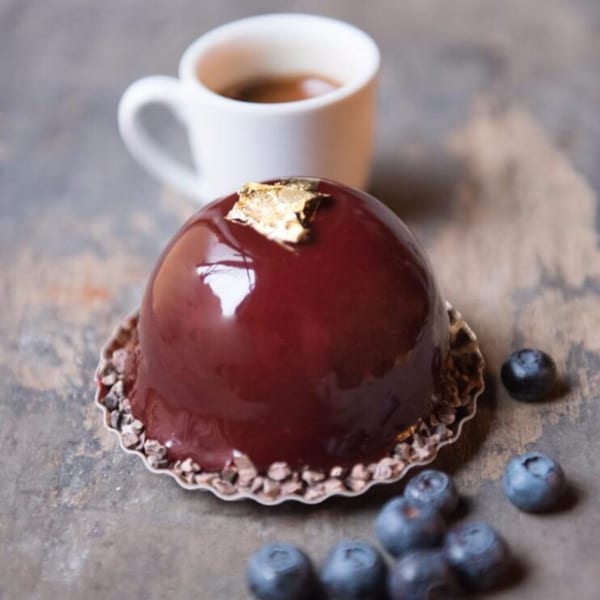 A shiny, dark red dome-shaped dessert with gold leaf, blueberries, and a cup of coffee.