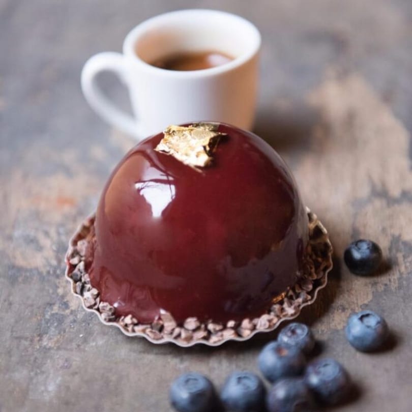 A shiny, dark red dome-shaped dessert with gold leaf, blueberries, and a cup of coffee.