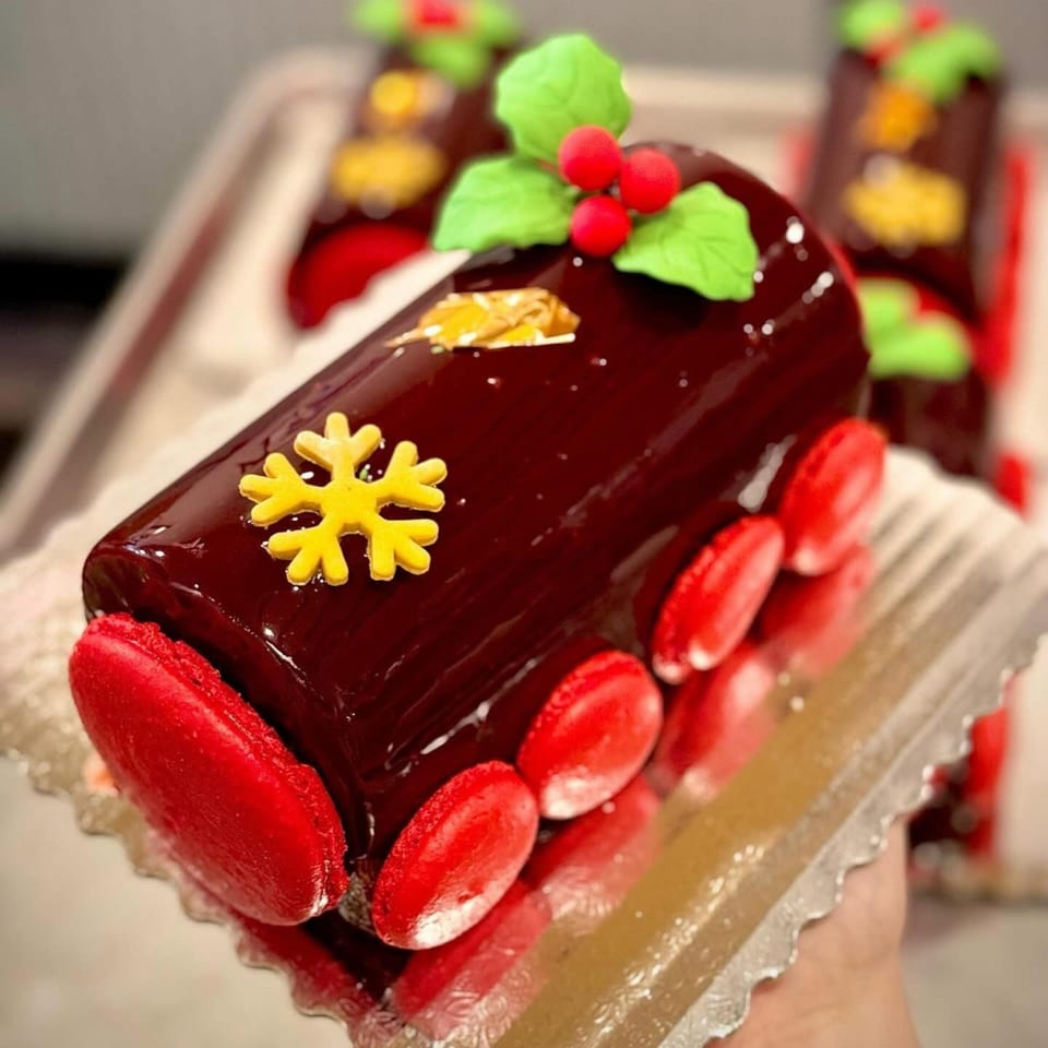 A festive chocolate Yule log cake with red macarons, a yellow snowflake, and holly decorations.